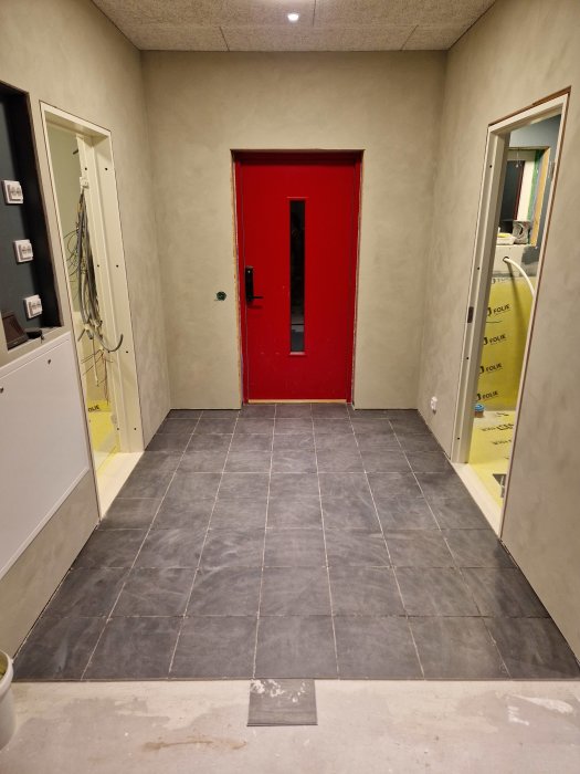 Ett rum under renovering med röd dörr, grått golv, oskyddade vägguttag och isolering synlig.