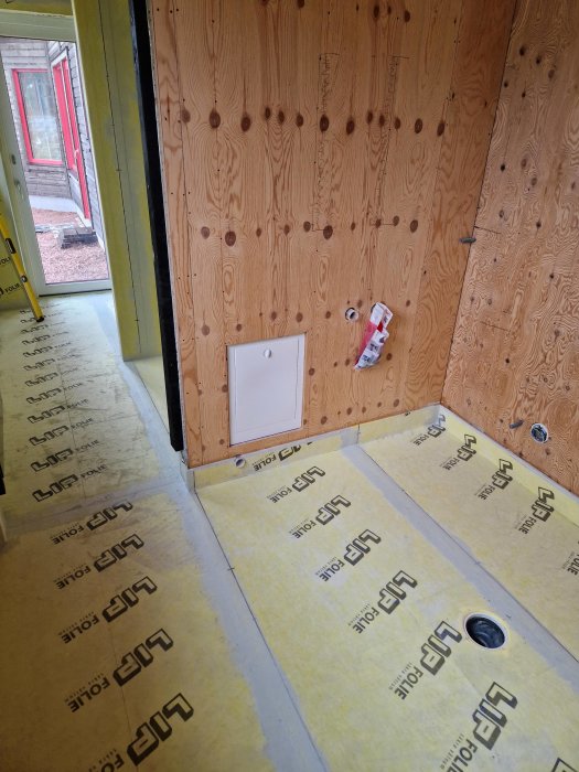 Inomhusbyggnadsarbete med synlig isolering, plywoodväggar och oavslutat eluttag, öppen dörr till utsidan.