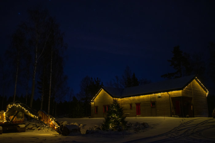 Trästuga med julbelysning, snö, stjärnklar himmel, vinternatt och dekorerad grävmaskin.