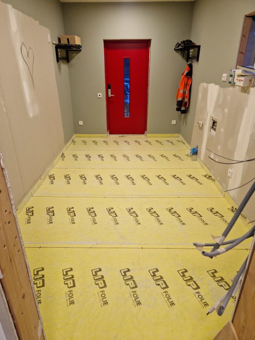Rum under renovering med röd dörr, isolerade golv, oavslutade väggar och arbetskläder.