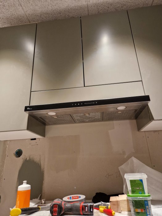 Kök under renovering, fläktkåpa, skåp, verktyg och byggmaterial synliga, oavslutade väggar, installation pågår.