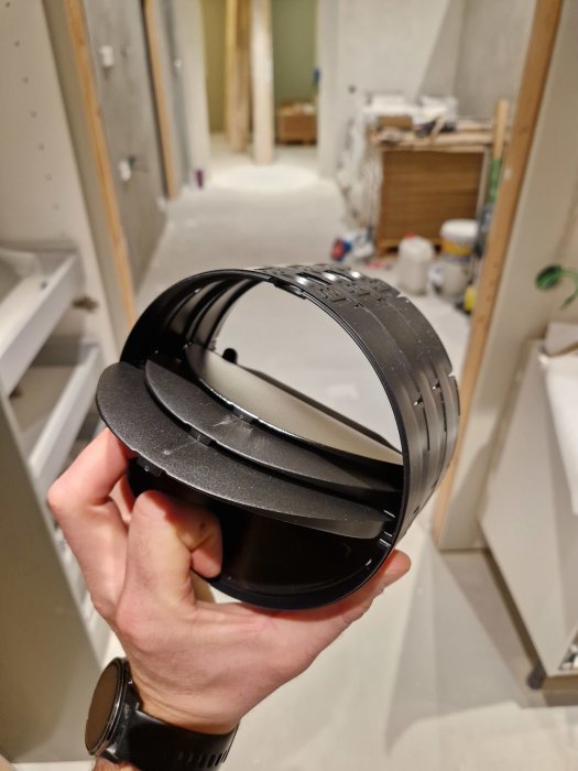 En hand håller ett svart ventilationsrör framför en orenoverad inomhusmiljö.