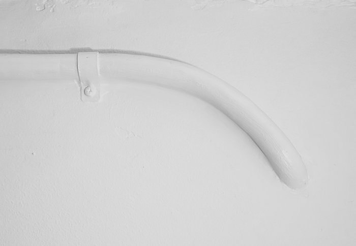 Monokrom bild på en vitmålad kabelklämma som håller en böjd kabel mot en vägg.
