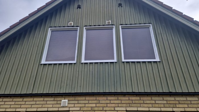 En fasad med tre fönster under ett sadeltak, persienner neddragna, gröna plåt- och gula tegelväggar.