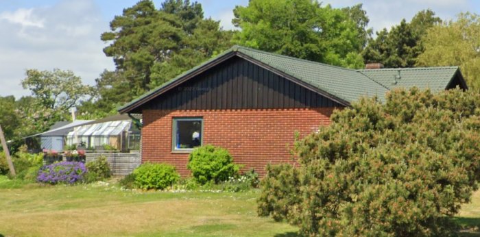 Ett rött tegelhus med mörkt tak, trädgård, växthus och gröna träd under en klar himmel.
