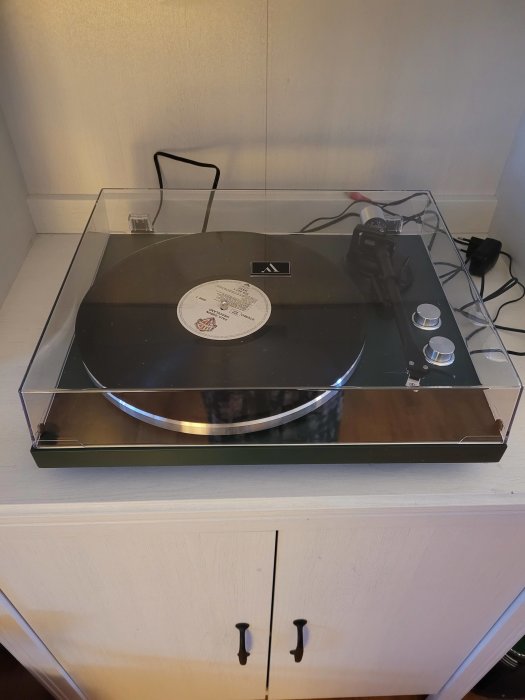 Skivspelare med vinylskiva, genomskinligt dammskydd, reglage, placerad på vit byrå.