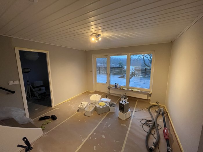 Ett rum under renovering, täckt golv, målarfärg, fönster med utsikt, verktyg och skor.