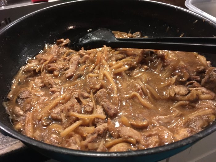 En stekpanna med en kött- och pastarätt, sås, stekspade i köksmiljö.