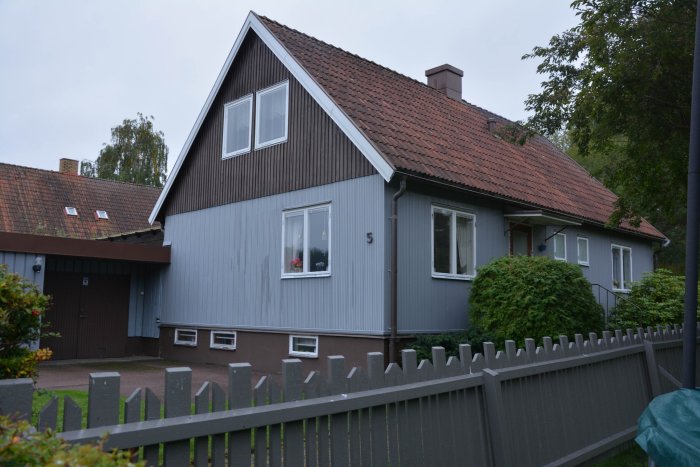 Ett grått hus med sadeltak och röda takpannor, omgiven av ett staket och grönska.