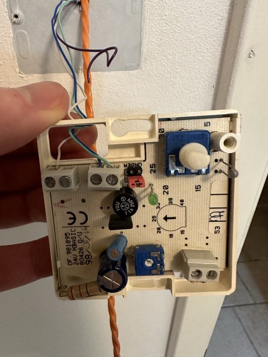 En demonterad termostat visas, med synliga komponenter, kretskort och trådar inne i en vägg.