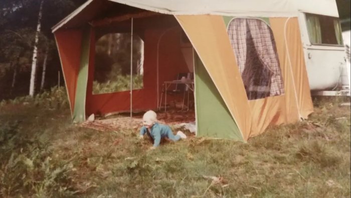 Ett litet barn kryper utanför en stor färgglad campingtält intill en husvagn i en skogsmiljö.