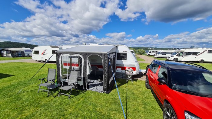 Campingplats med husvagnar, förtält, bilar, grönt gräs, delvis molnig himmel.