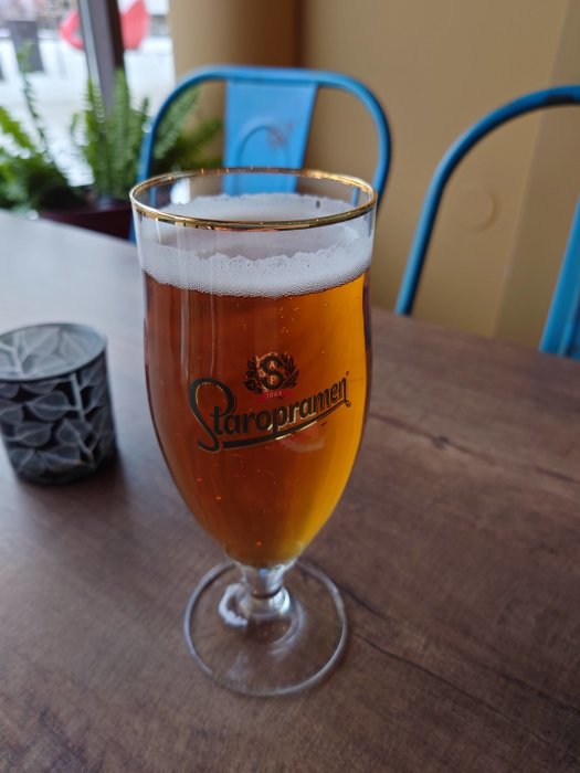 Ölglas med skum, märkt "Staropramen", träbord, blå stol, utomhusmiljö.