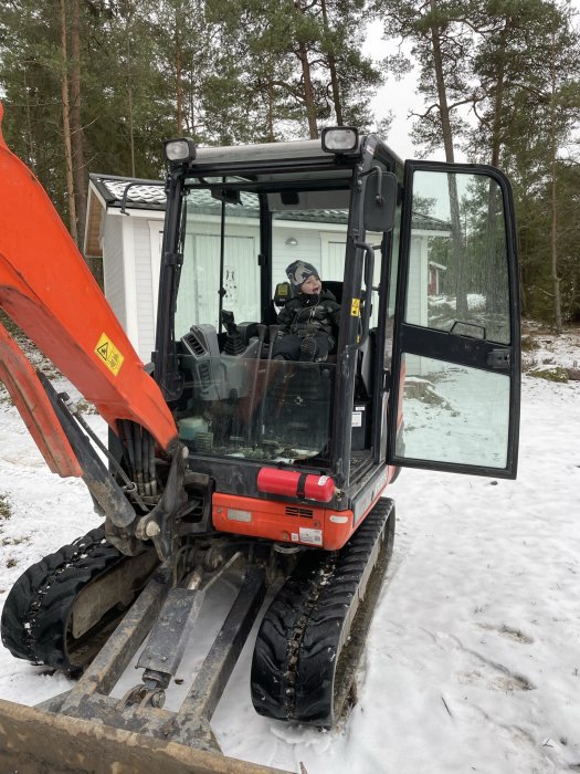 Barn i grävmaskinshytt, snötäckt mark, träd i bakgrunden, bandgrävare, lek eller lärande.