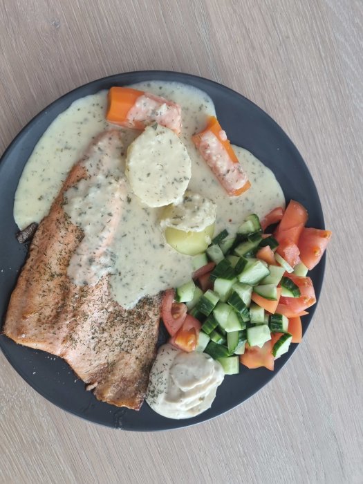 Grillad lax med dillsås, kokt potatis, morot, rå gurka och tomatsallad på en mörk tallrik.