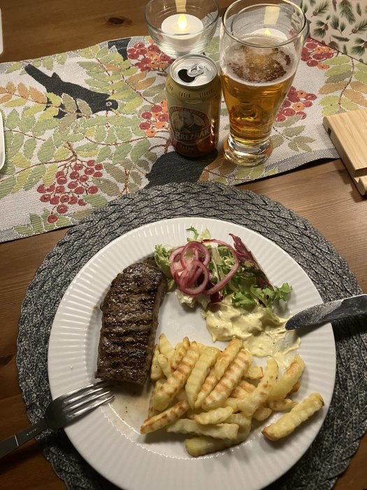 En måltid med biff, pommes frites och sallad, öl och tända ljus på ett bord.