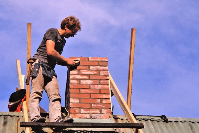 Man arbetar på ett tak, bygger skorsten, klar himmel, arbetskläder, verktygsbälte, säkerhetsrisk.