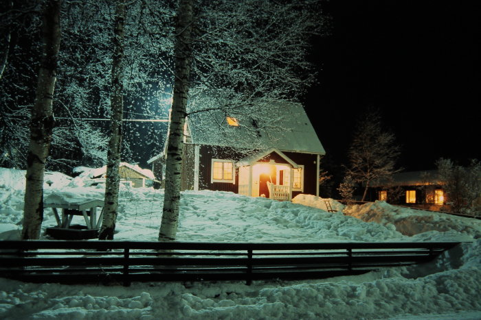 Vinter, snö, stuga med belysning, träd, kväll, fridfullt, picknickbord, gärdsgård, kallt.