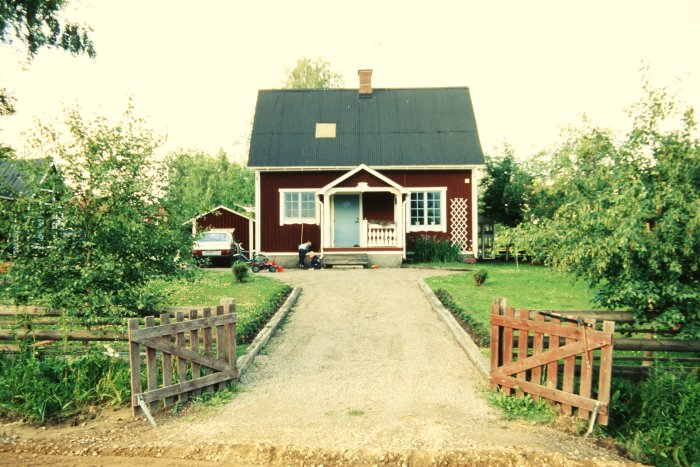 Rött hus med vita knutar, grusgång, grönska, person, leksak, bil, trästaket, sommar, lugn, hemtrevligt.