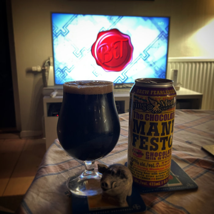 Ölglas med mörkt öl, burk, TV-skärm i bakgrunden, hipposammetsfigur, duk på bordet.