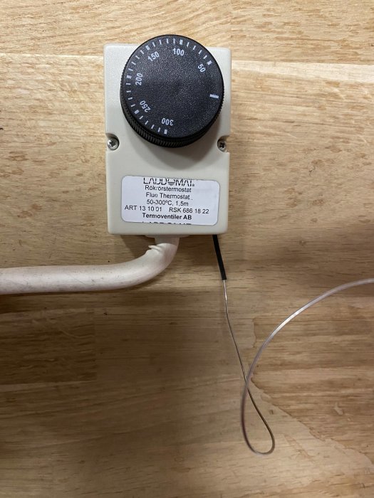 Väggmonterad röktermostat med justerbar ratt och sensorledning i en inomhusmiljö på trävägg.