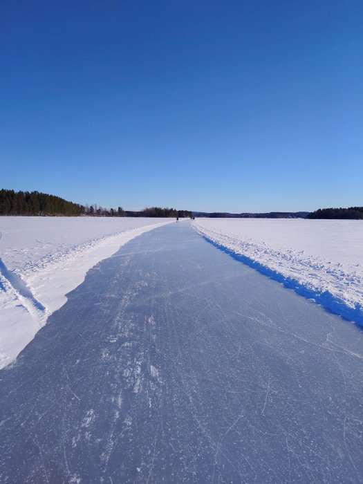 Klarblå himmel, isbelagd sjö, personer långt borta, skog i horisonten, snöbanker vid sidorna.