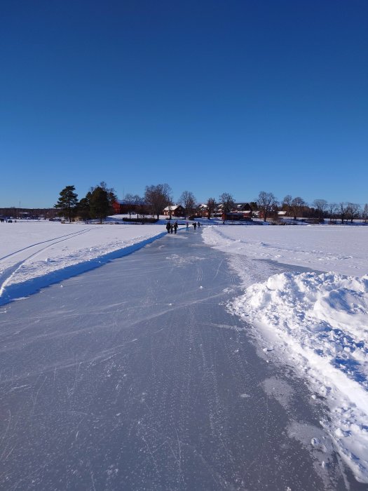 Klart väder, personer åker skridskor på en frusen sjö, snötäckt landskap, träd och hus i bakgrunden.