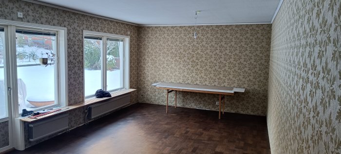 Föråldrat rum med blommig tapet, fönster mot snötäckt landskap, enkelt bord, mörkt laminatgolv, radiatorer under fönstren.