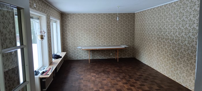 Tomt rum med blommigt tapet, trägolv, fönster, bord, radiator och lite saker på fönsterbrädan.