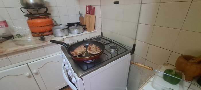 Kött steks i stekpanna på gasolspis i ett kök med kakelväggar och köksredskap.