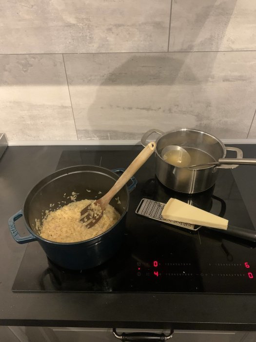 Induktionshäll med två kastruller, träredskap, risotto in progress, osthyvel, modernt kök.