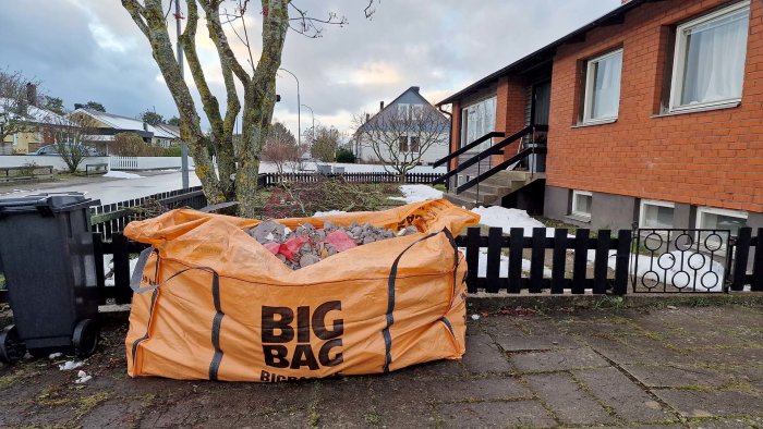 Stor orange säck fylld med byggavfall framför tegelhus, grå himmel, bar trädstam, svart soptunna, snötäckt mark.