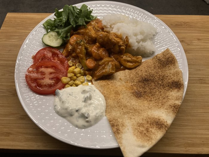 En tallrik med curry, ris, naanbröd, sallad, tomat, gurka och raita.