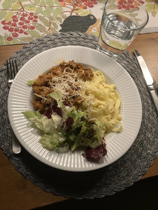 En tallrik med pasta, köttsås, riven ost och sallad vid ett matbord. Glas med vatten. Mönstrad duk.