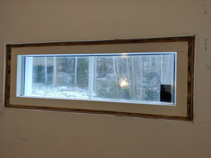 Långsmalt fönster visar vinterskog och snö från ett ouppfärdigt inomhusutrymme.