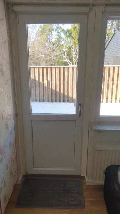 Utsikt från ett rum genom en glasdörr, visar snötäckt altan och träd, med tapetserade väggar inuti.