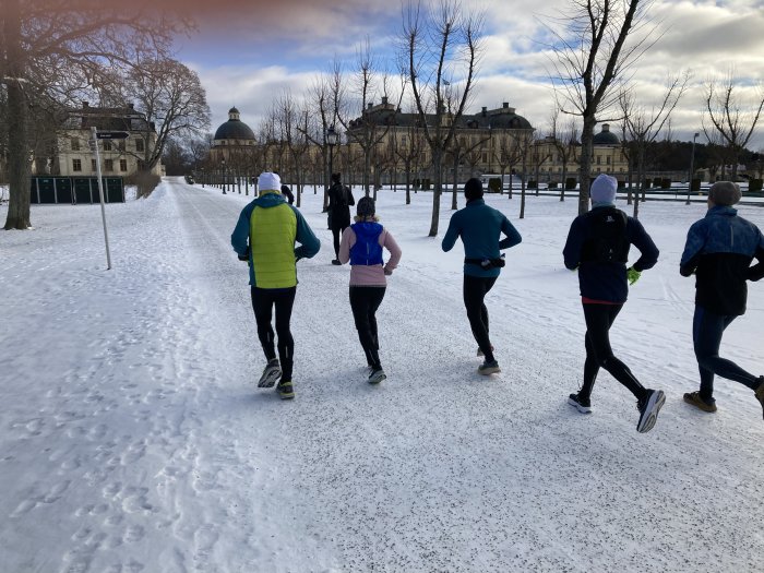 Grupp människor joggar tillsammans på snötäckt väg vid historisk byggnad i solljus.