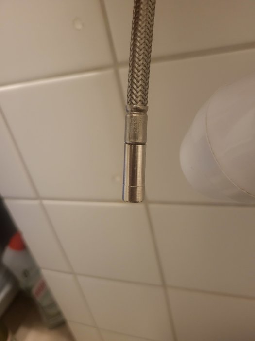 Metallic flexibel rörända nära vita plattor och duschdel, möjligen en duschslang eller vattenanslutning.