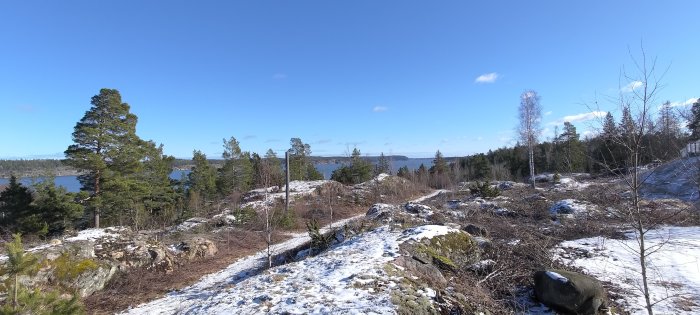 Skogsklädd kulle, snöfläckar, klippor, blå himmel, sjö i bakgrunden, solig vinterdag.