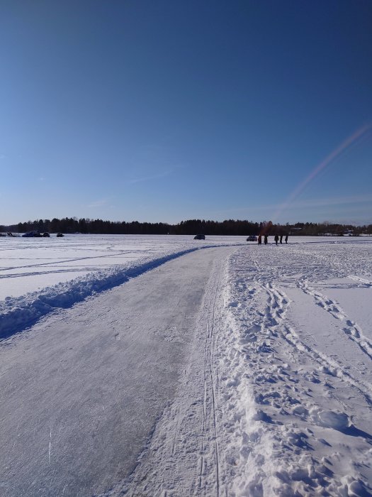 Snötäckt landskap, isbelagd väg, personer i fjärran, klarblå himmel, soligt, vinterdag.