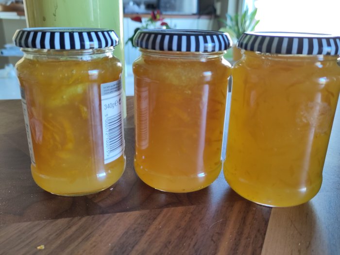Tre burkar med marmelad eller sylt, randiga lock, på ett träbord. Transparent orange innehåll.