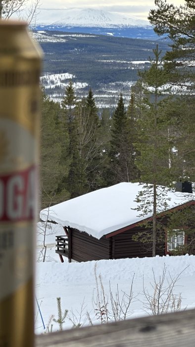 Utsikt över snötäckt landskap med fjäll, träd och stuga, förgrundsoskärpa på ölburk.