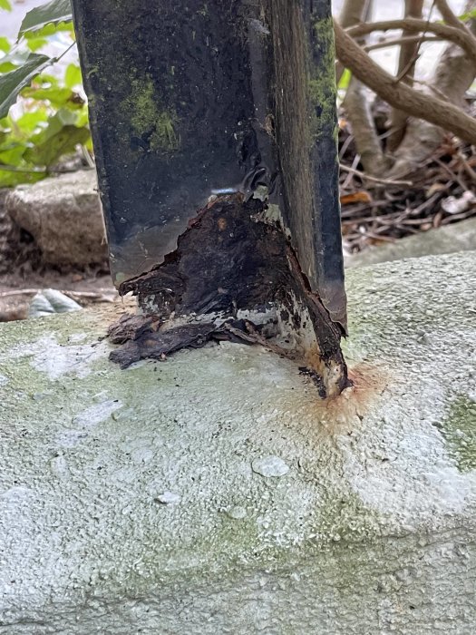 Metallstolpe med rostskada vid basen, på en cementyta utomhus, med vegetationsbakgrund.