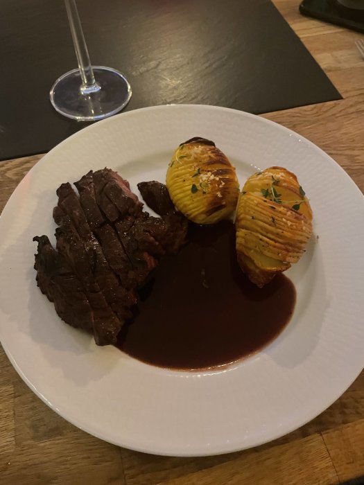 Grillat entrecôte med hasselbackspotatis och rödvinssås på en vit tallrik. Eleganta, enkla presentationer. Glas i bakgrunden.