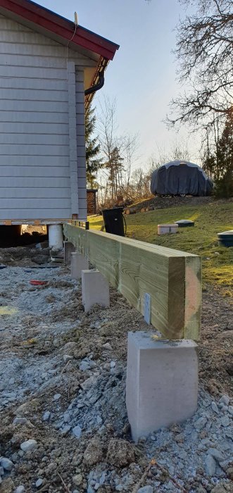 Huslyft, grå betongpelare, träbjälkar, grusig mark, nedgrävda rör, skymmande träd i bakgrunden, blå himmel, skymningen.