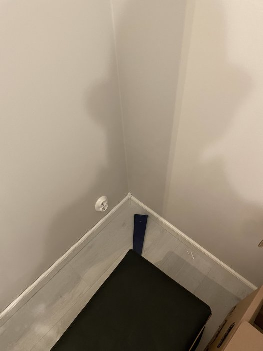 Hörn av ett rum med grått golv, vita väggar och svart matta. Skuggspel skapar illusion av person.