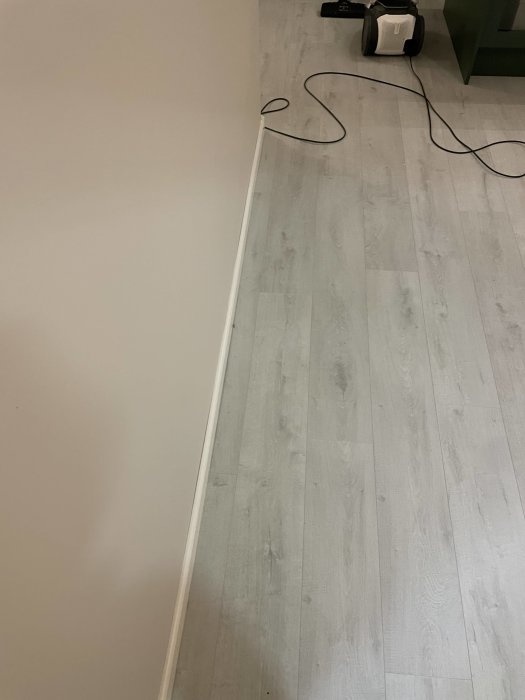 Ett inomhusrum med ljusgrått laminatgolv, en vit vägg och en dammsugare i bakgrunden.