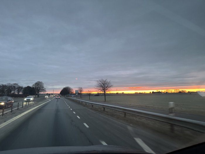 Motorväg, trafik, gryning, molnigt, barträd, färgad himmel, bilkörning, öppet landskap, vägmarkeringar.