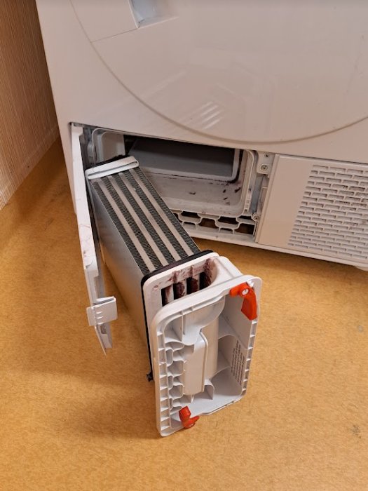 Öppen torktumlare med utdragen kondensor, står på ett beige golv mot vit vägg.