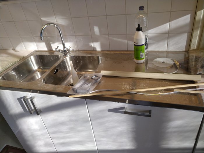 Köksskåp och diskbänk med rörigt byggmaterial, verktyg och rengöringsmedel i solljus. Renovering eller installation pågår.